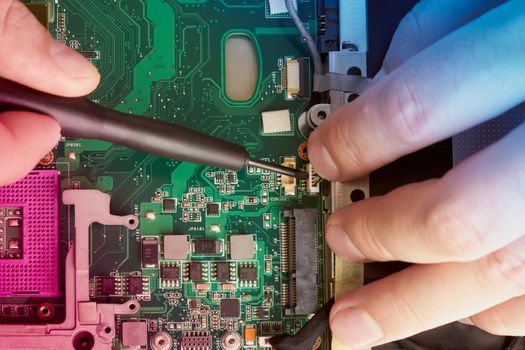 Man repairs computer. A service engineer in a shirt repairs a laptop, at a white Desk against a white wall. Computer motherboard background with blur neon multicolor glow. Hands close up.