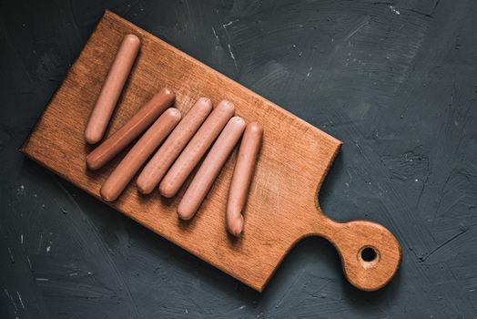 Boiled fried sausages sausages lie on a wooden kitchen board scratched against a dark concrete background.