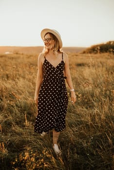 A fair-skinned blonde girl in a dress with polka dots in a hat walks through the meadow among the tall grass in the summer at sunset.