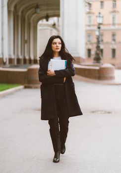 Beautiful serious smart girl brunette student holding notebooks and textbooks, goes walking at University on street of St. Petersburg. A charming woman with long dark hair is studying at a course