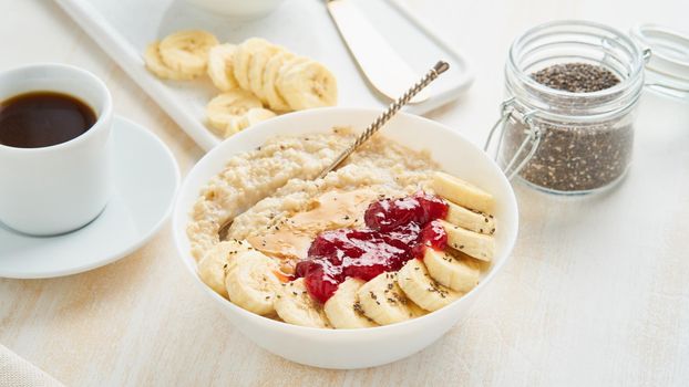 Close up of oatmeal porridge, healthy vegan diet breakfast with strawberry jam, peanut butter, banana, chia on white wooden light background. Side view
