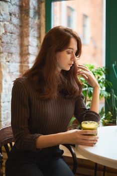 Beautiful serious stylish fashionable smart girl sits by the window in a cafe and drinking healthy yellow smoothie or latte vegan. Charming thoughtful woman with long dark brown hair.