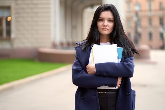 Beautiful serious smart girl brunette student holding notebooks and textbooks, stands at University on street of St. Petersburg. A charming woman with long dark hair is studying at a course