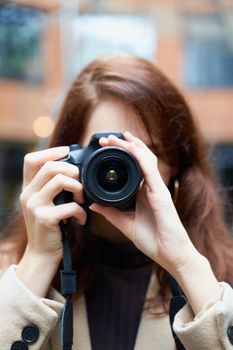 Selective focuson lens. Beautiful stylish fashionable girl holds camera in her hands and takes pictures. Woman photographer with long dark hair in a city, urban shoot, vertical. unrecognizable person