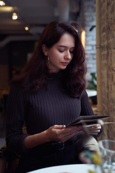 Beautiful serious stylish fashionable smart girl sit in cafe in loft style on lunch. Ready to place an order, studying menu. Charming thoughtful woman with long dark brown hair, vertical, moody