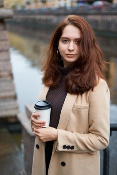 Beautiful serious stylish fashionable girl holding cup of coffee in hands walking street of St. Petersburg in city center. Charming thoughtful woman with long dark hair looks at camera, vertical