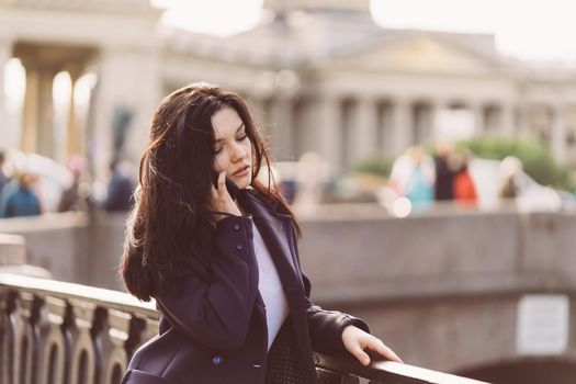 Woman with long hair talking on phone. Autumn or winter, girl in outdoor. Beautiful intelligent brunette in street of Saint-Petersburg in city center near bridge, copy space