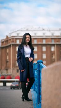 Charming thoughtful fashionably dressed woman with long dark hair travels through Europe, standing in city center of St. Petersburg. A beautiful girl wanders alone through autumn streets, vertical