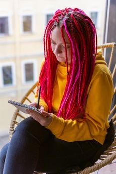 Girl artist, illustrator draws in notebook, makes sketches. Close-up of woman with long pink dreadlocks in informal setting, in casual comfortable clothes sitting in a cafe, vertical