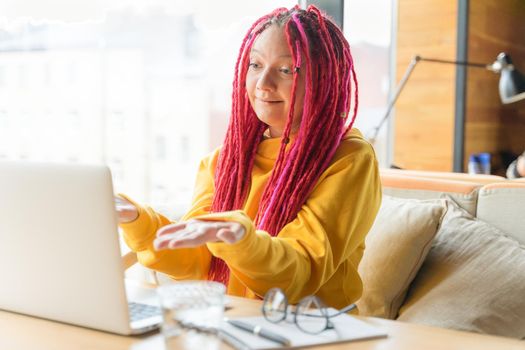 Woman looks at a laptop and is surprised, indignant, spread her hands. Girl with long pink hair, creative designer, freelancer. Concept of difficulties in working with virtual online communication