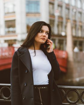 Woman with long hair talking on phone. Autumn or winter, girl in outdoor. Beautiful intelligent brunette in street of Saint-Petersburg in a city center near bridge, copy space, vertical