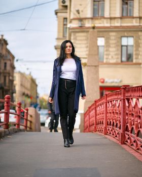 Charming thoughtful fashionably dressed woman with long dark hair travels through Europe, walking in the city center of St. Petersburg. A beautiful girl wanders alone through the autumn streets