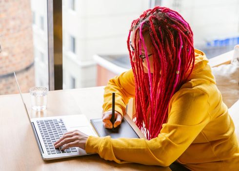 Girl designer, illustrator draws on a tablet, looks into a laptop. The concept of the remote work of a creative hobby, freelance. Woman with long pink hair, bright appearance.