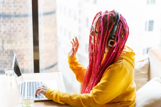 Woman with headphones looks at laptop and waves her hand. Live chat, stream. Girl with long pink hair, designer, creative specialist, freelancer, blogger. Good luck concept, positive online