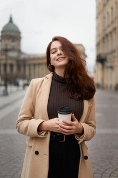 Beautiful serious stylish fashionable smart girl holding cup of coffee in hands and smiles, goes walking down street of St. Petersburg in city center. Charming thoughtful woman with long dark hair