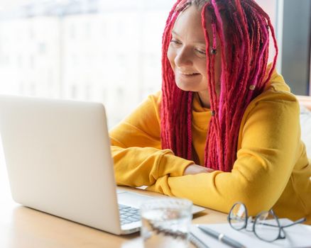 A woman is looking at a laptop and smiling. Live chat, stream. Girl with long pink hair, designer, creative specialist, freelancer, blogger. Good luck concept, positive online communication.
