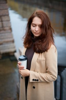 Beautiful serious stylish fashionable girl holding cup of coffee in hands walking street of St. Petersburg in a city center. Charming thoughtful woman with long dark hair looks down, vertical