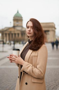 Beautiful serious stylish fashionable smart girl holding cup of coffee in hands goes walking down street of St. Petersburg in city center. Charming thoughtful woman with long dark hair, vertical