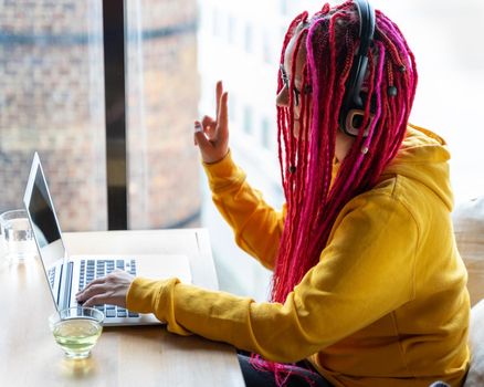 The woman in headphones looks at the laptop and shows a victory sign with her hand. Live chat, stream. Girl with long pink hair, freelancer, blogger. Good luck concept, positive communication.