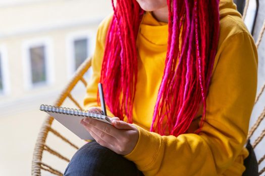 Unrecognizable person, girl artist, illustrator draws in notebook, makes sketch. A woman with long pink dreadlocks in informal setting, in casual comfortable clothes is sitting in a cafe
