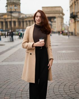 Beautiful serious stylish fashionable smart girl holding cup of coffee in hands goes walking down street of St. Petersburg in city center. Charming thoughtful woman with long dark hair, vertical