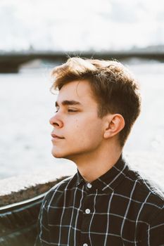 Portrait of a young guy, brunette with dark hair, Caucasian. Close - up of a guy, boy, man looking past camera. Outdoor in city by water