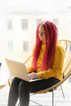 Digital nomad concept. Girl freelancer remotely working on a laptop in a cafe, coworking. Woman with long pink dreadlocks in an informal setting, in casual comfortable clothes sitting at chair, vertical