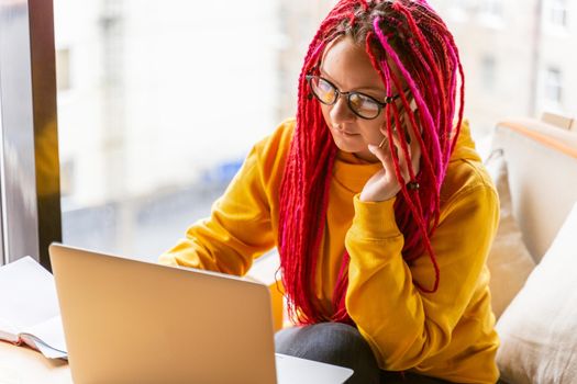 Concept of digital nomad. Girl freelancer talking on mobile phone, working remotely on laptop in cafe, coworking. Woman with long pink dreadlocks in informal setting, in a casual clothes
