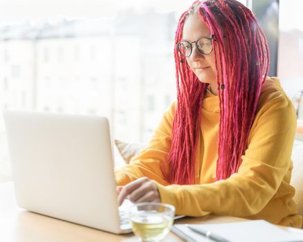 A woman is looking at a laptop and is typing on a keyboard. Girl with long pink hair, designer, creative specialist, freelancer, journalist. Digital Nomad Concept