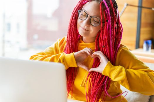 Woman looks at laptop and shows heart shape sign with her palms. Live chat, stream. Girl with long pink hair, designer, creative specialist, freelancer, blogger. Good luck concept. Close up