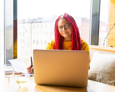 Girl with pink long hair, dreadlocks works at computer in cafe. Concept of remote work, freelance, digital nomad . Designer is planning design project. Desk with laptop, notepad in coworking space