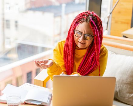Woman looks at a laptop and is surprised, indignant, spread her hands. Girl with long pink hair, creative designer, freelancer. Concept of difficulties in working with virtual online communication