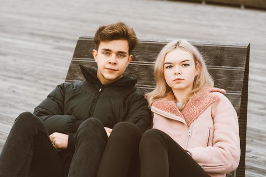Loving teenagers are sitting on park bench in autumn, looking straight ahead. Teenage love concept. Portrait of cute brunette guy in dark jacket, beautiful young blonde girl in light pink jacket.