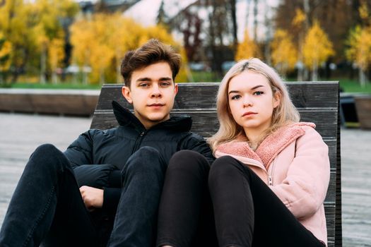 Loving teenagers are sitting on park bench in autumn, looking straight ahead. Teenage love concept. Outdoor portrait of cute brunette guy in dark jacket, beautiful young blonde girl in light pink jacket.