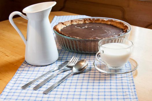 Chocolate cheesecake in a glass baking dish. Near glass cup of milk, knife, fork, spoon and milk jug