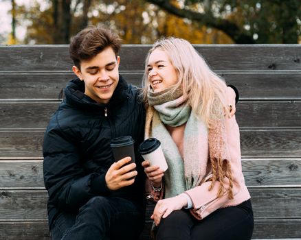 Lovely brunette guy and pretty girl blonde drink coffee and chat on date, laugh. Loving teenagers are sitting on park bench. Teen Love Concept. They are happy, smiling, laughing. Evening