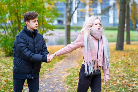 Two teenagers in love in quarrel. The blonde girl takes offense at boy, the guy holds her hand, asks the girl to stay and not leave. Concept of difficulties in adolescent relationships