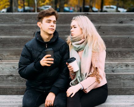 Lovely brunette guy and pretty girl blonde drink coffee and chat on a date. Loving teenagers are sitting on park bench. Teen Love Concept. They are happy, smiling, laughing