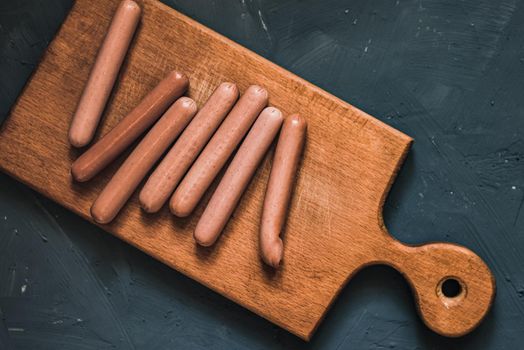 Boiled fried sausages sausages lie on a wooden kitchen board scratched against a dark concrete background.