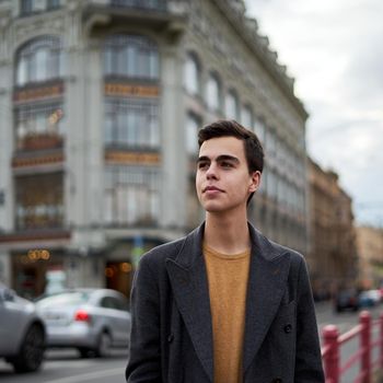 Handsome stylish fashionable man, brunette in an elegant gray coat, stands on street in historical center of St. Petersburg. Young man with dark hair, thick eyebrows.