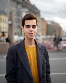 Handsome stylish fashionable man, brunette in elegant gray coat, stands on the street in historical center of St. Petersburg. Young man with dark hair, thick eyebrows, vertical