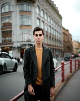 Handsome stylish fashionable man, brunette in elegant gray coat, stands on the street in historical center of St. Petersburg. Young man with dark hair, thick eyebrows, vertical