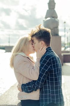 The boy looks tenderly at the girl and wants to kiss. A young couple stands embracing. The concept of teenage love and first kiss, sincere feelings of man and woman. The city, the waterfront. Vertical