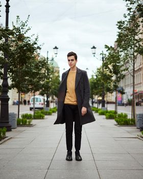 Handsome stylish fashionable man, brunette in an elegant gray coat, stands on street in historical center of St. Petersburg. Young man with dark hair, thick eyebrows, vertical