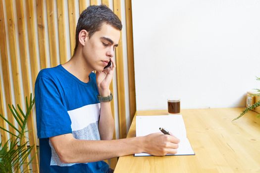 Young man writing information to notepad and speaking mobile phone, teenager in casual clothes plans a schedule, copy space
