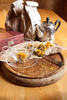 A glass cup, teespoon, kettle and dried marigold flowers on a wooden board.