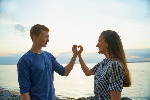 teen couple showing heart with hands on the beach at the sunset