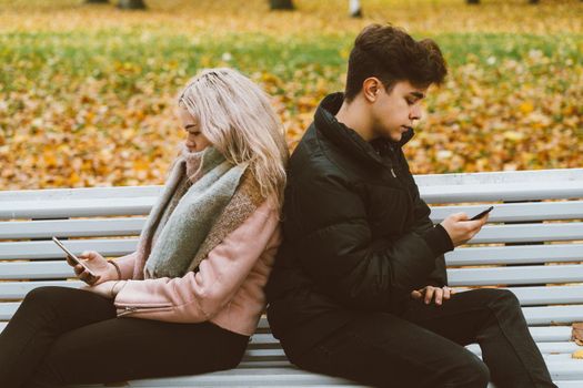 Loving teenagers on date look at mobile phones, sit on park bench in the fall. Immersion in virtual world, social networks. Concept of teenage love, Smombie. Teen dating, top view