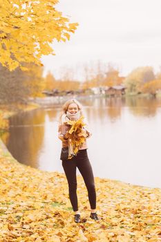 Beautiful young girl throws up autumn leaves. Attractive young woman is resting, fooling around, playing with leaves in the park, vertical