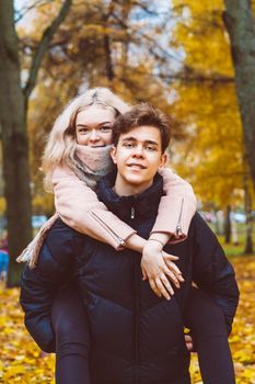 A pretty blonde girl is sitting on her back with a charming brunette man. Loving teens are happy, smiling, looking at the camera. Concept of teenage love walking outdoors